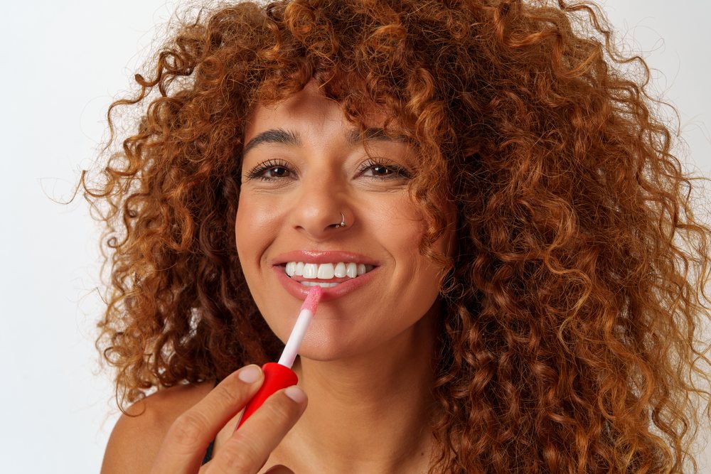 Hispanic woman applying lip gloss with curly red hair