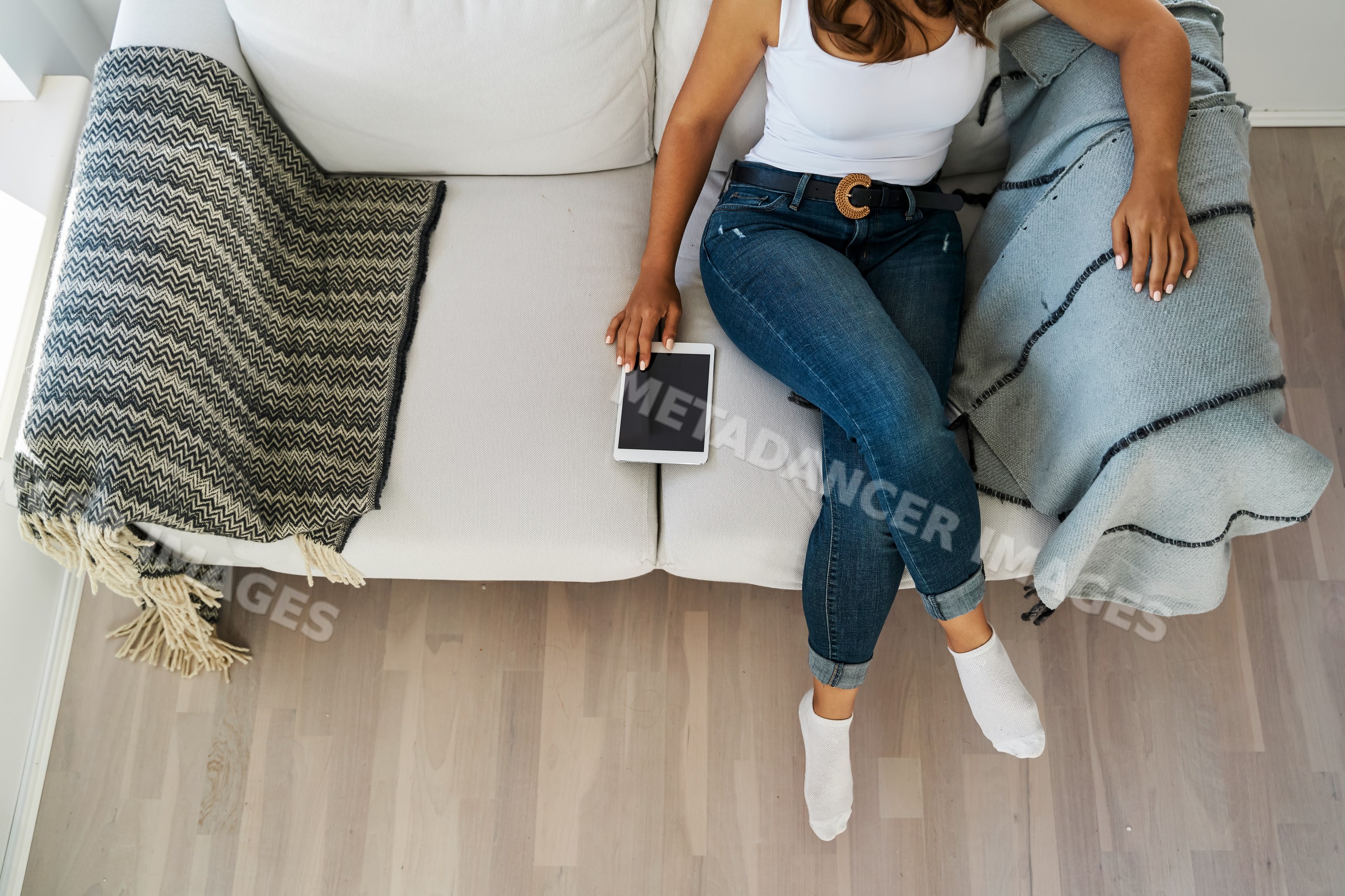 A white woman sits on a sofa holding a tablet indoors