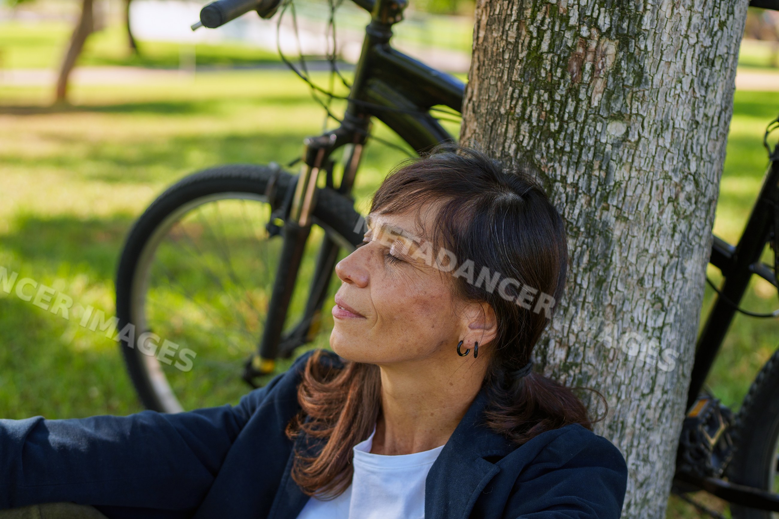 Hispanic woman relaxing against tree with bicycle in park in Málaga