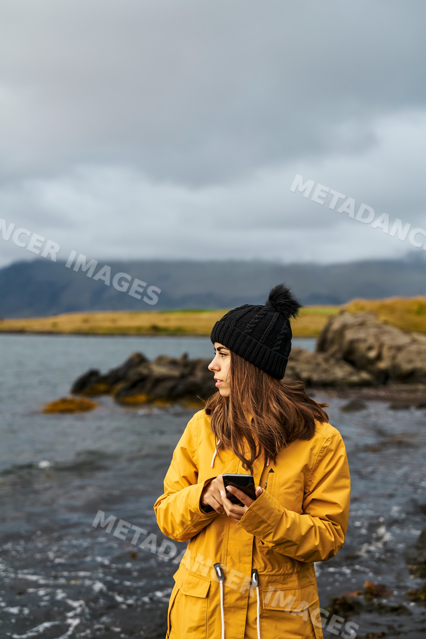 A white woman in a yellow jacket uses a smartphone by the shore
