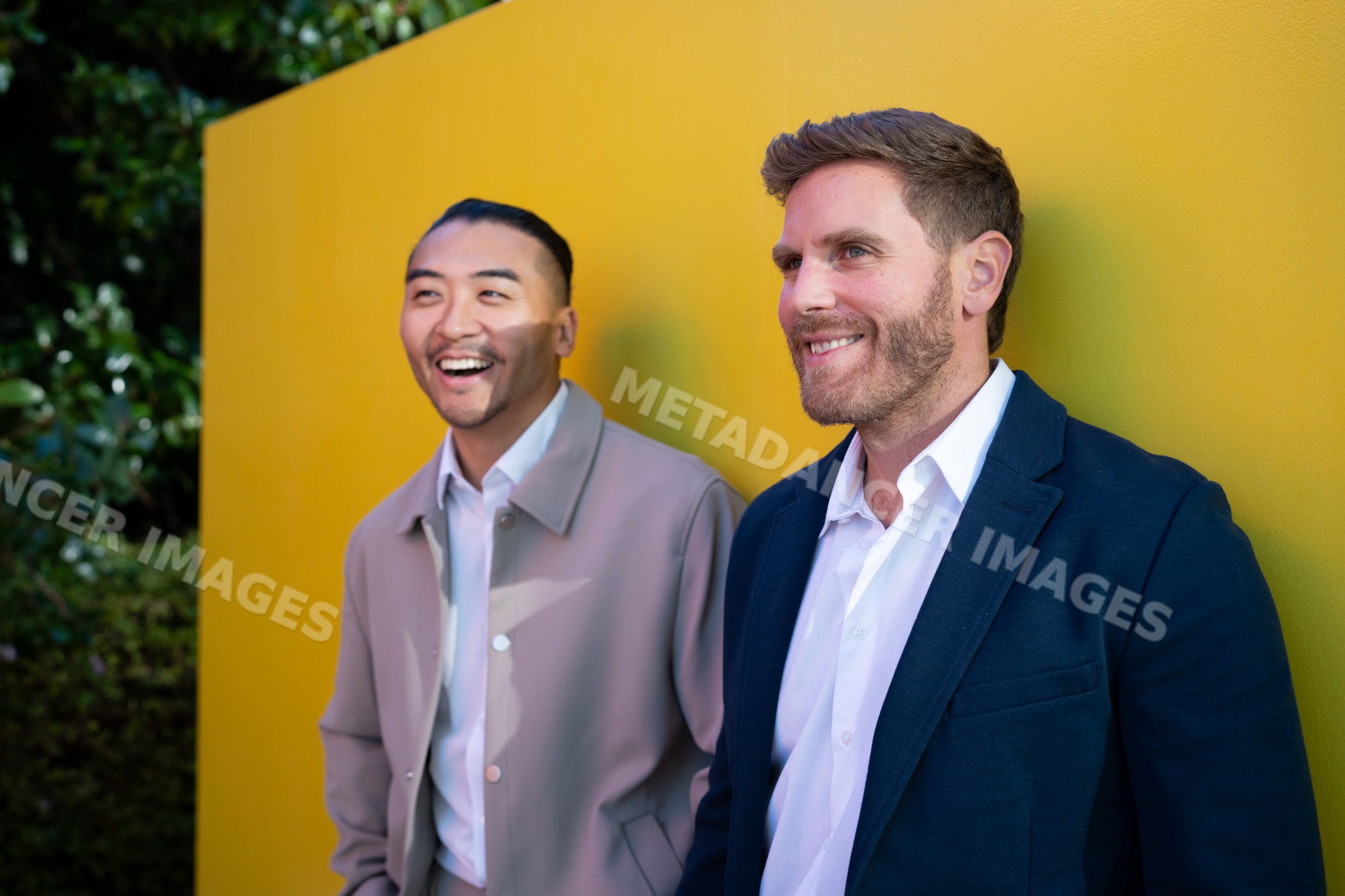 Two white men smiling outdoors against a yellow wall