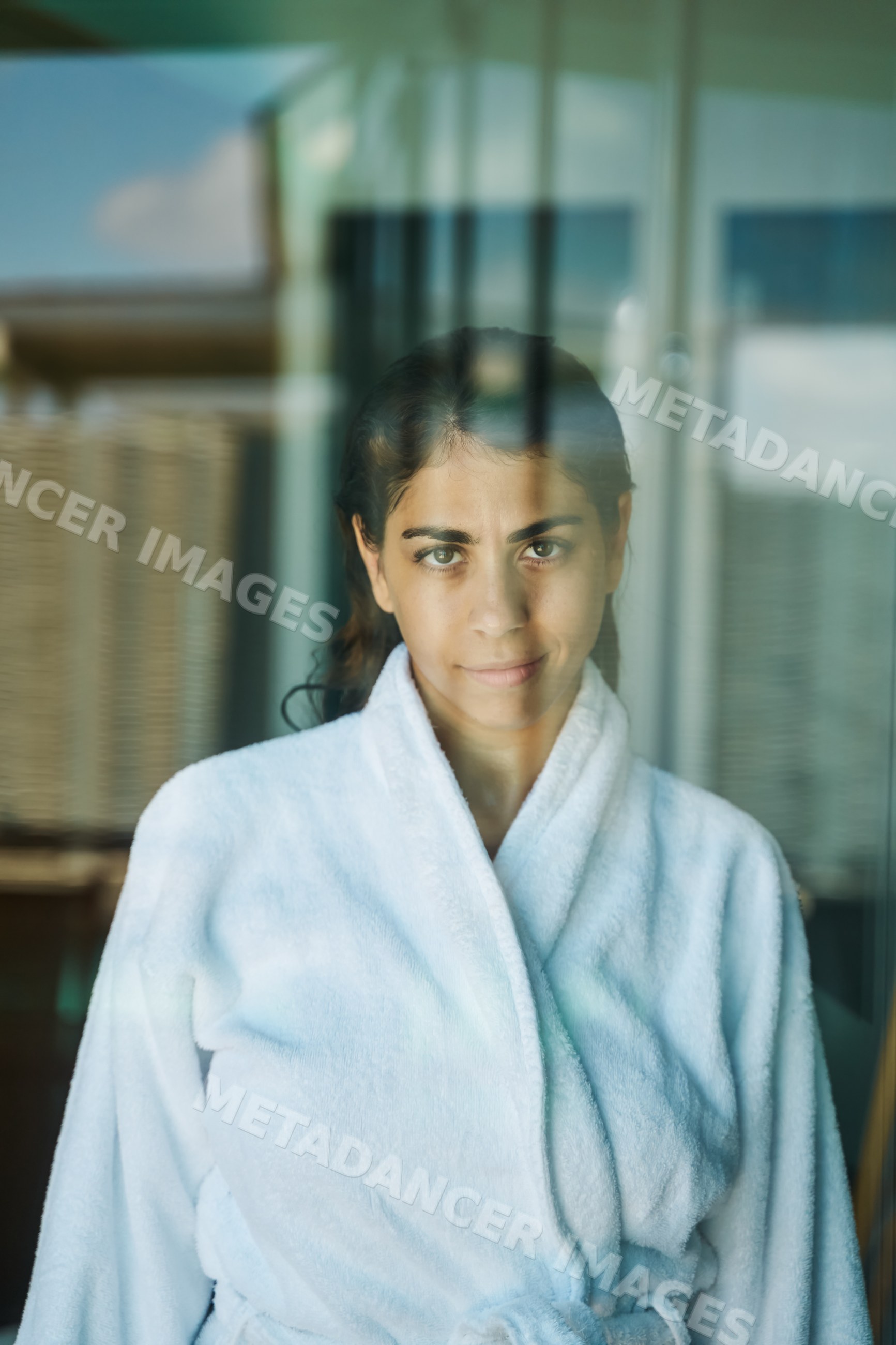 A woman in a white bathrobe relaxes indoors through a glass window