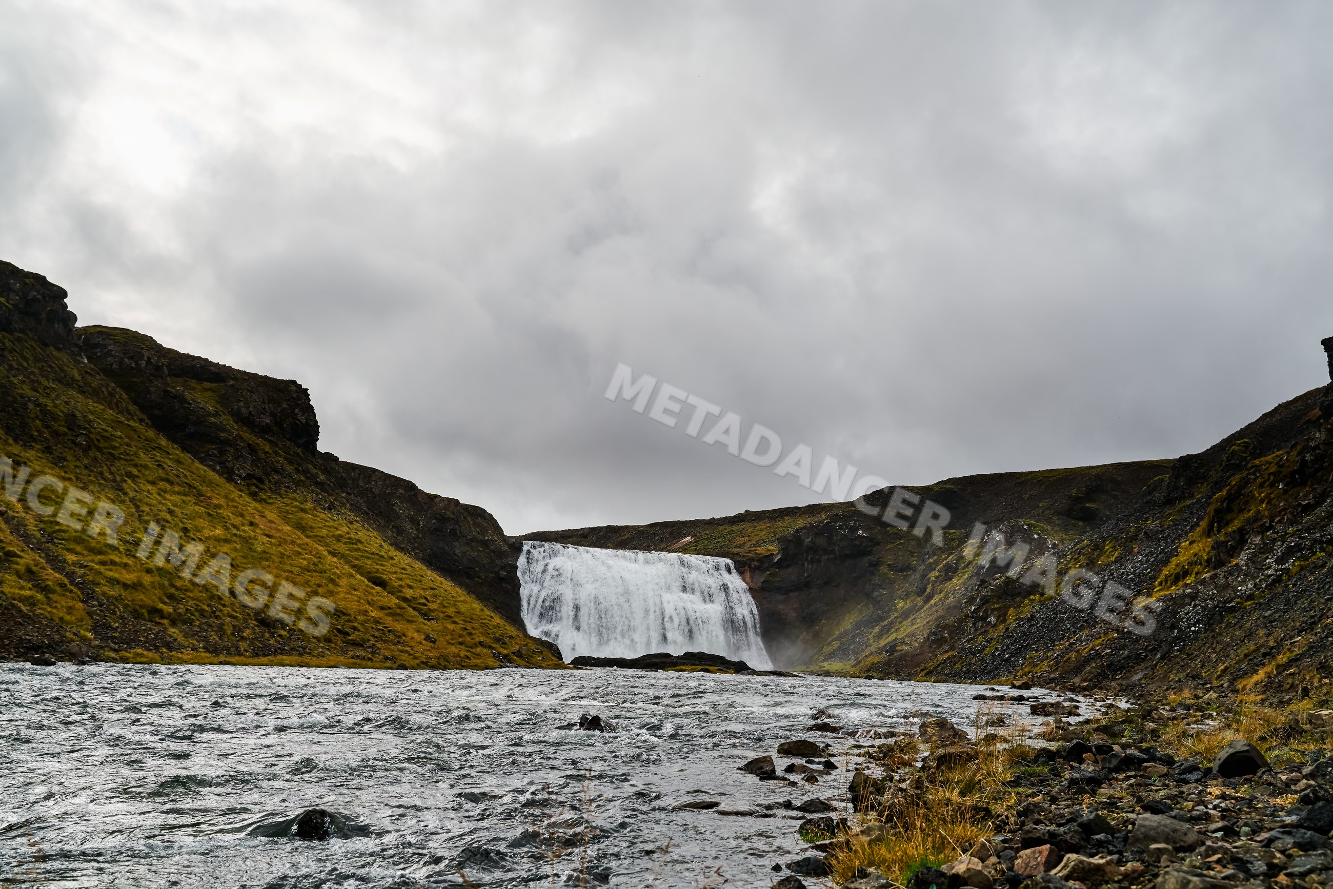 A waterfall flows into a river amid rocky hills and cloudy skies