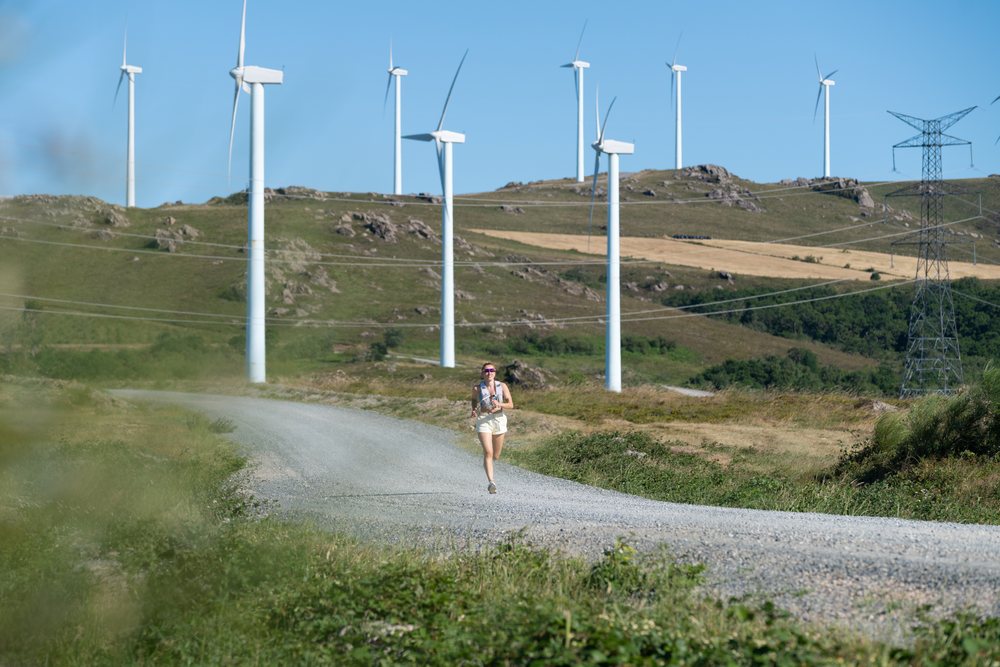 A white woman running on a gravel trail near wind turbines in Galicia