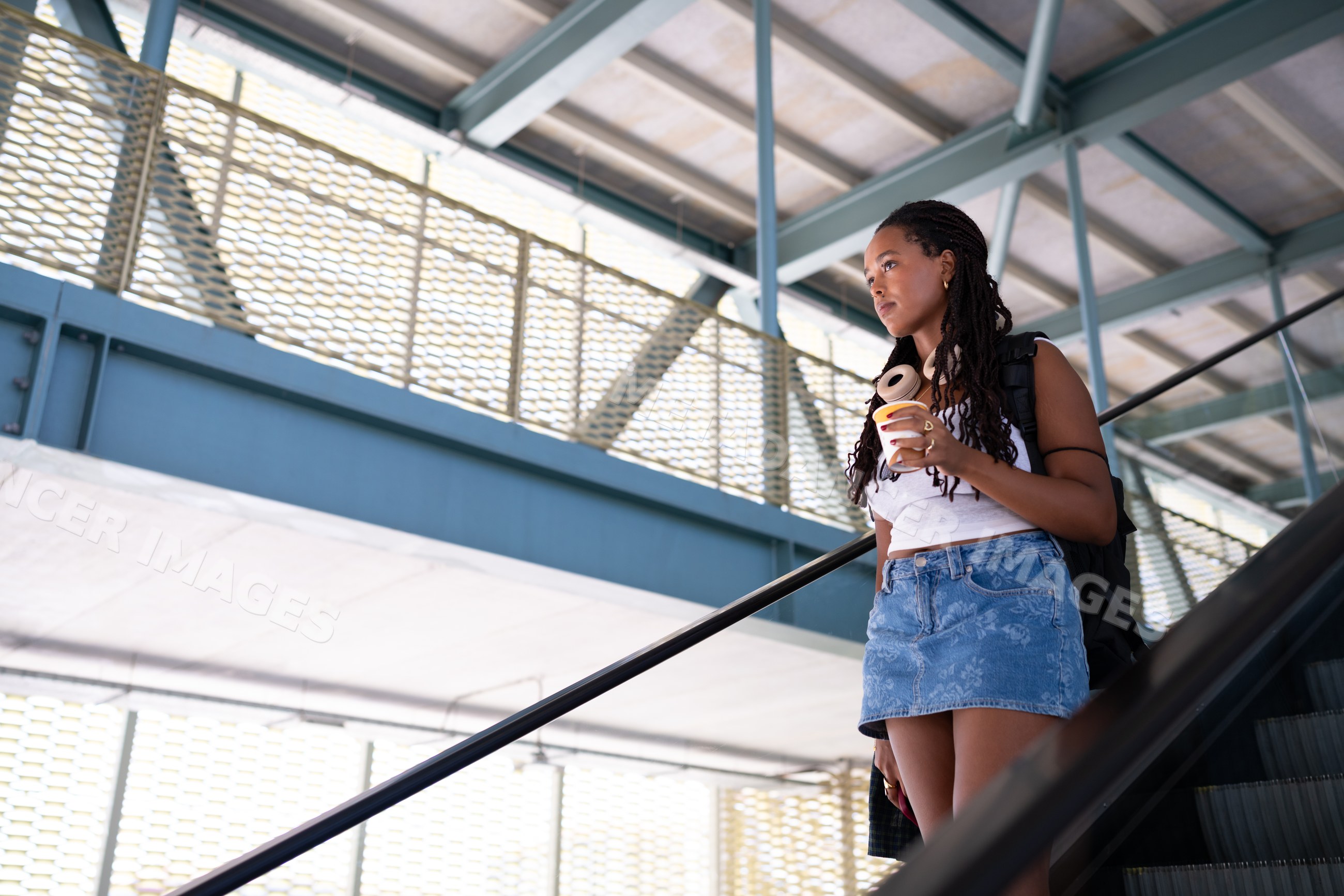 A young African American woman stands on an escalator holding coffee