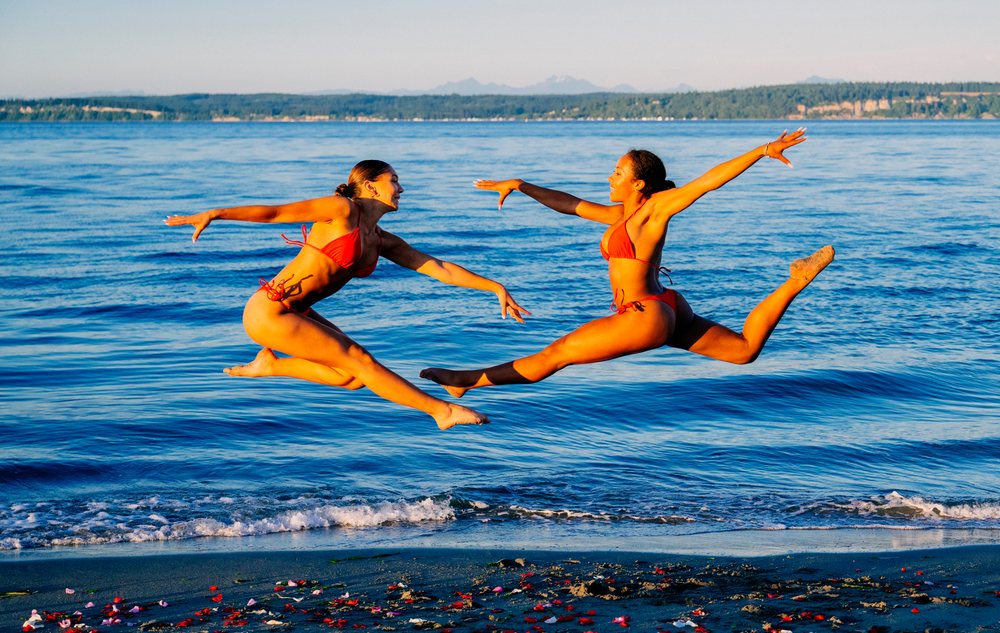 Two women jump in unison on a beach shoreline in WA, USA