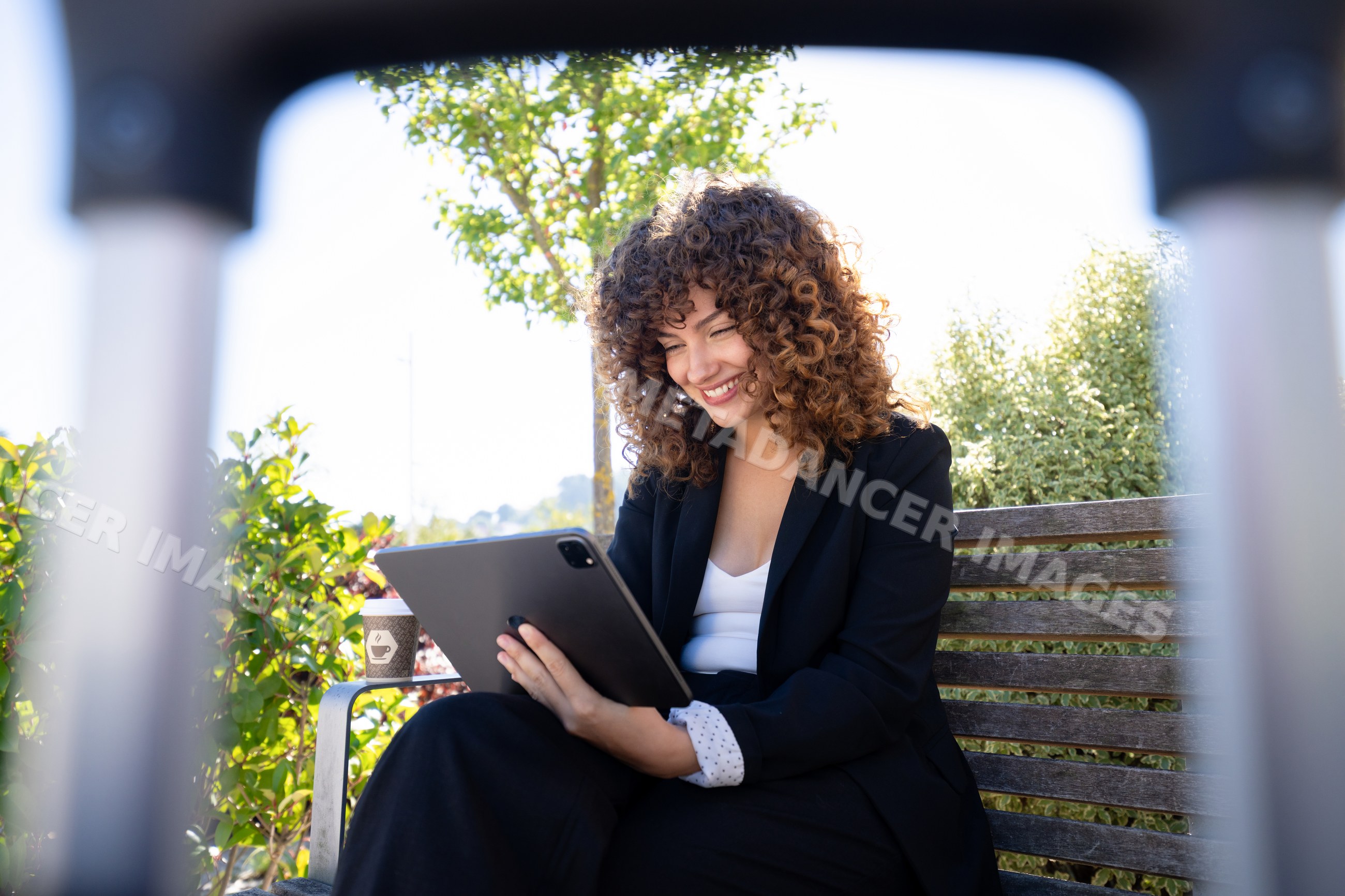 White woman smiling on outdoor bench using tablet with coffee cup