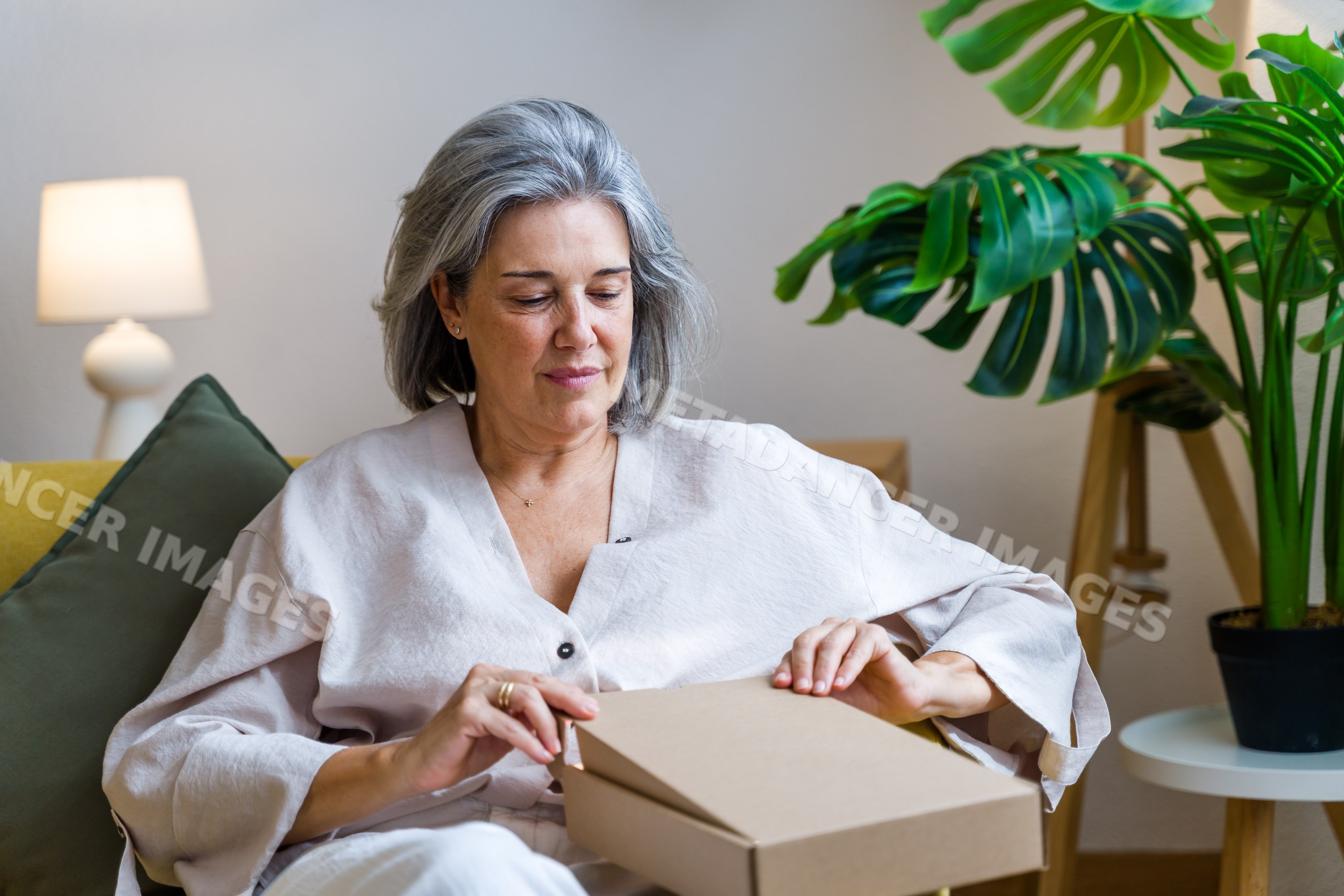 Mature white woman opening cardboard box indoors on couch with plants