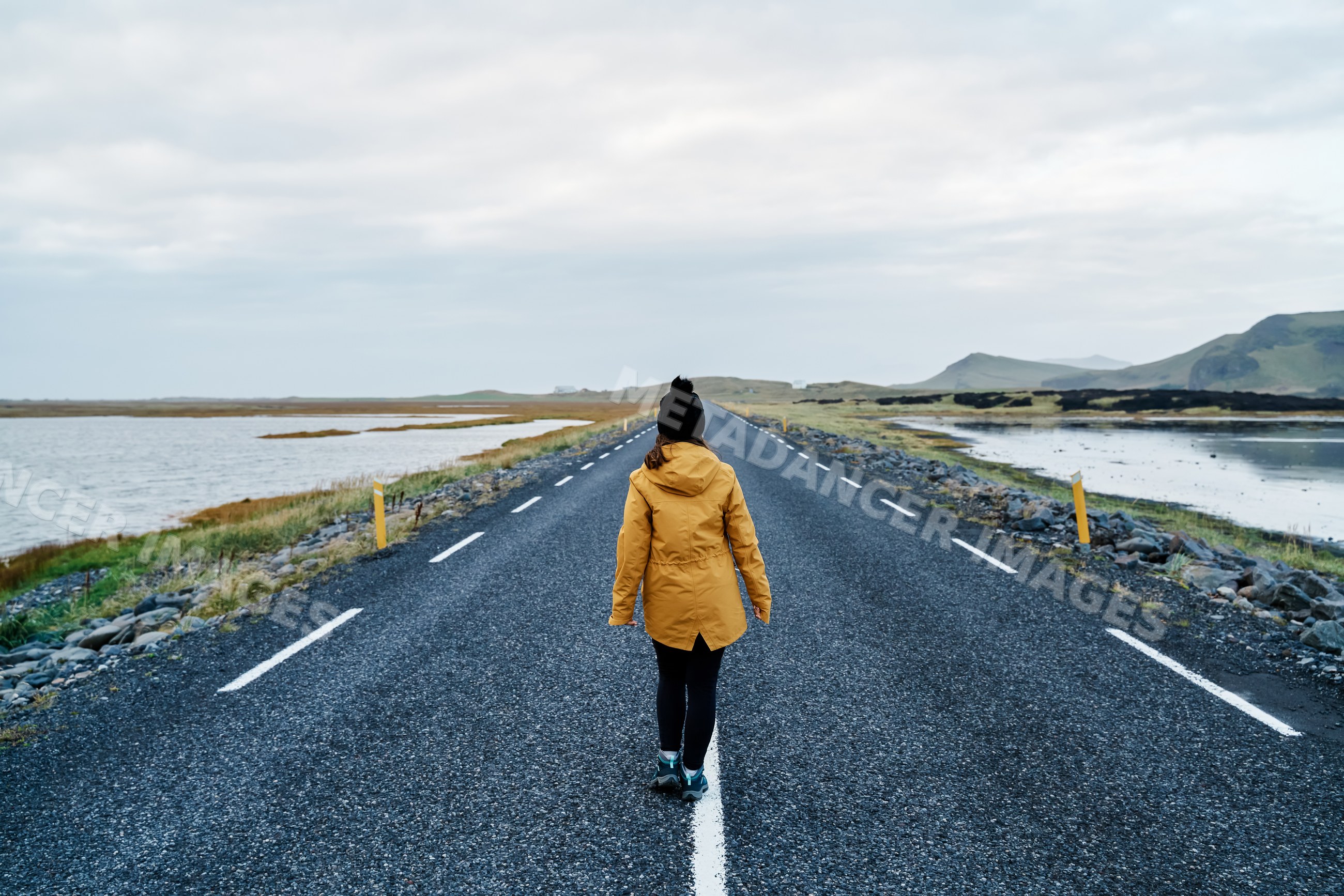 A white woman walks on an empty road surrounded by water and hills