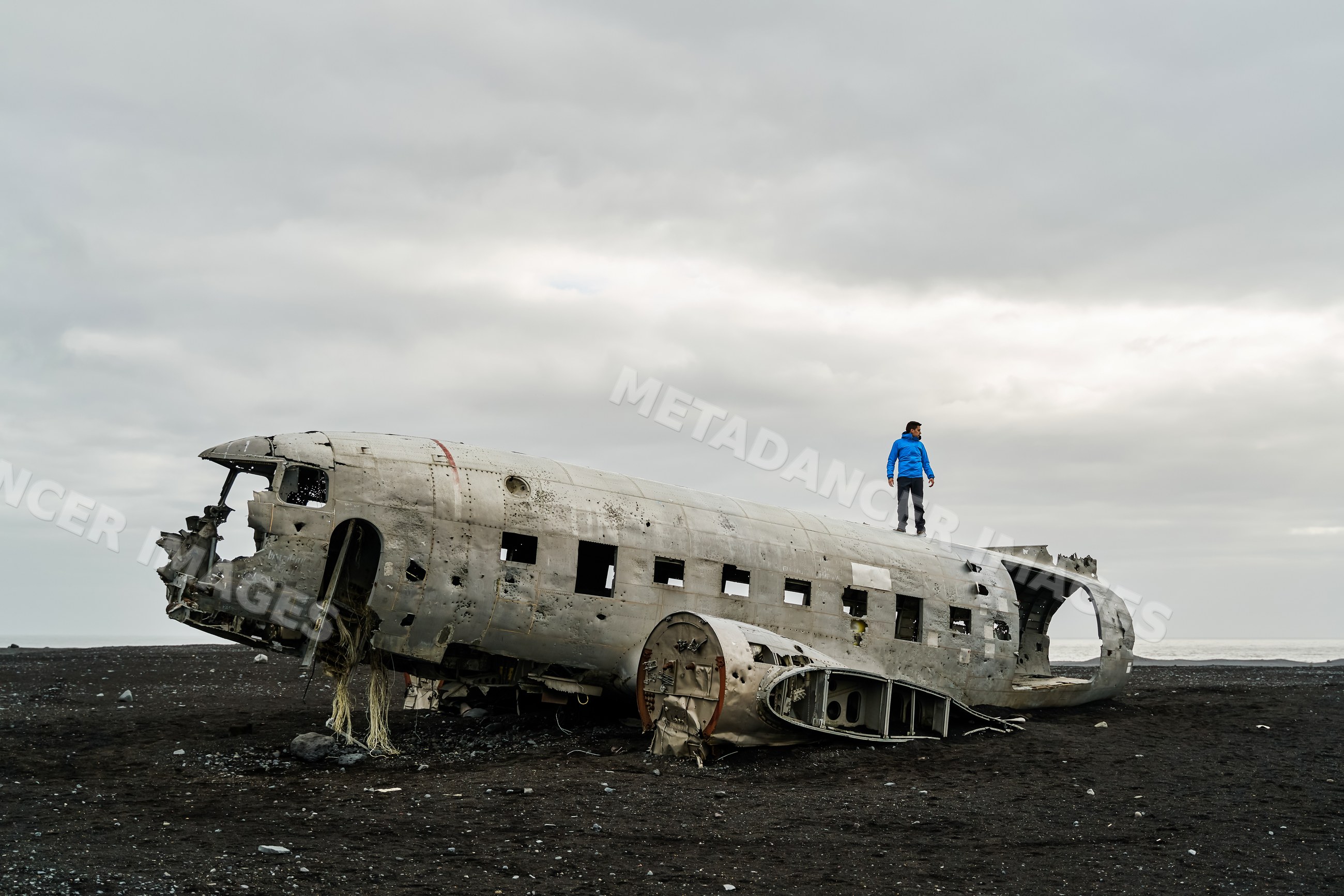 A white man explores the wreck of a crashed plane on a black sand
