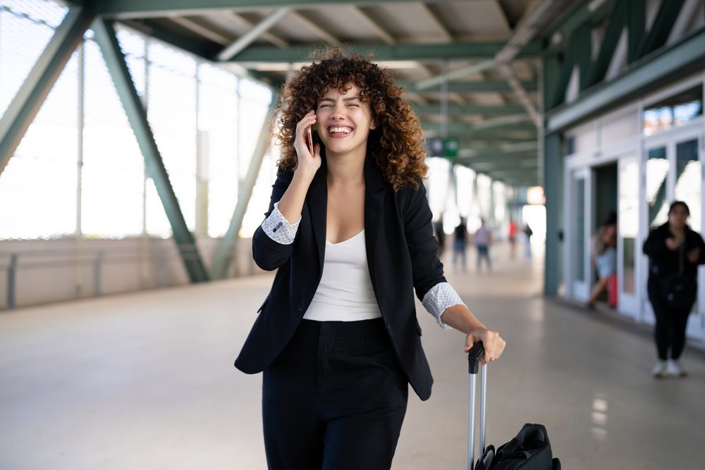White woman talking on phone walking with luggage in airport corridor