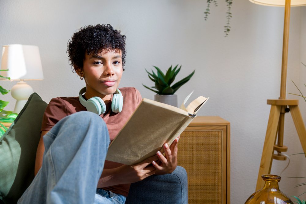 Young African American woman reading book at home with headphones