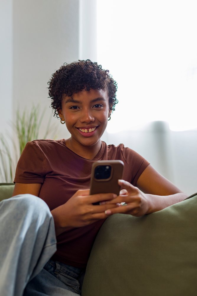 Young African American woman happily using smartphone sitting on couch