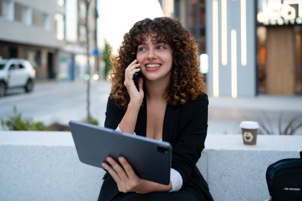 Young white woman holding tablet and talking on phone outdoors