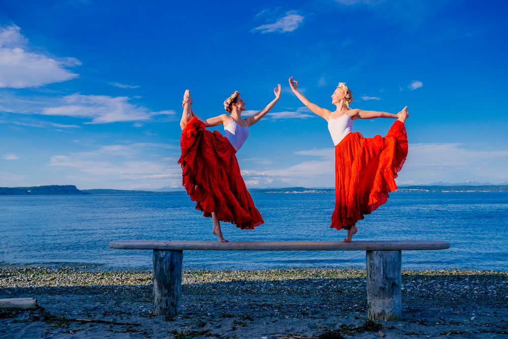 Two white women practice partner yoga acrobatics on a bench by the sea