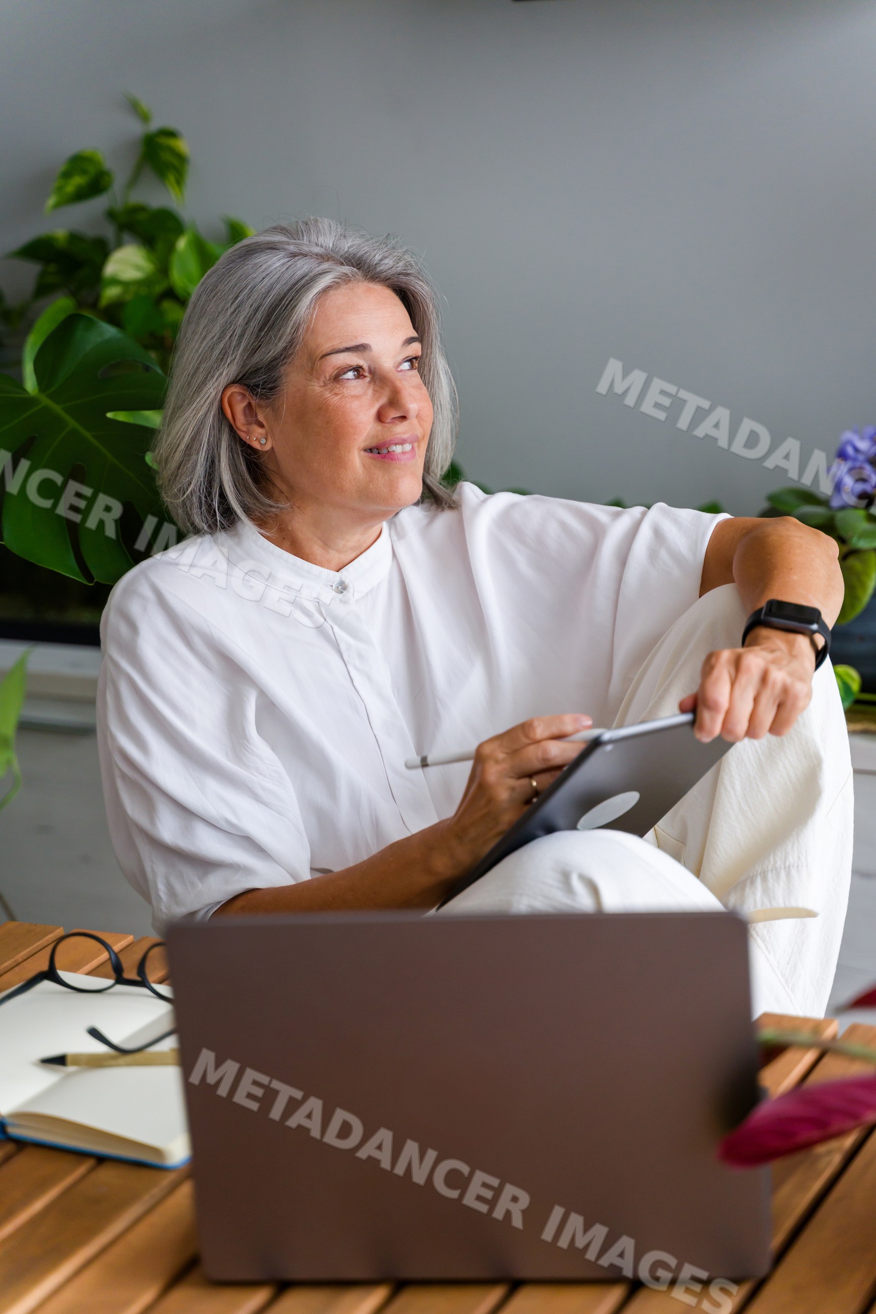 Mature white woman using digital tablet in home workspace with plants