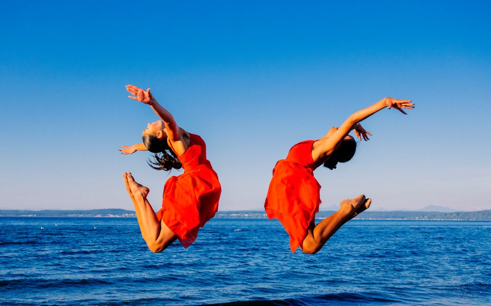 Two women in red dresses jump in acrobatic poses by the ocean shore