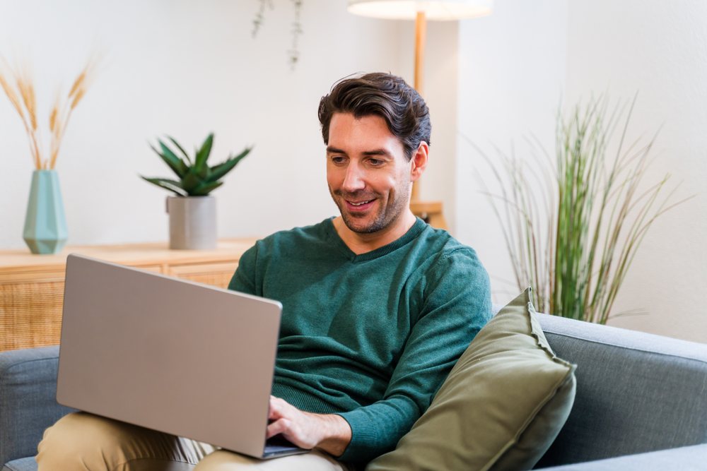 White man sitting on gray couch using laptop in modern living room