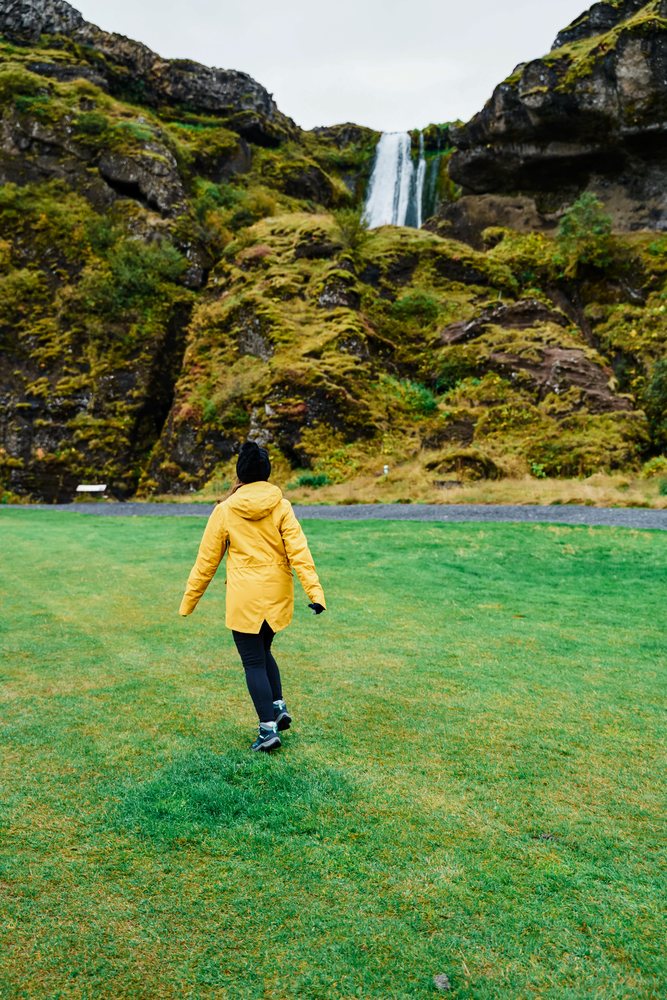 A white woman walks through grassy field toward a waterfall