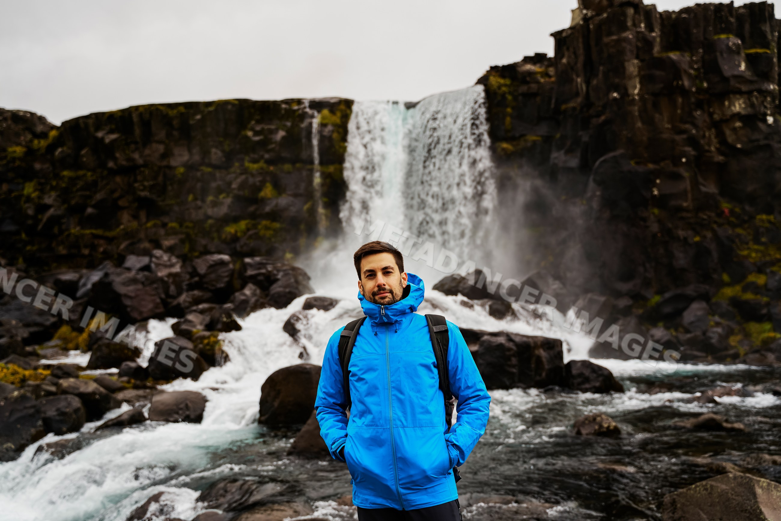 A white man stands near a waterfall in Iceland wearing a blue jacket