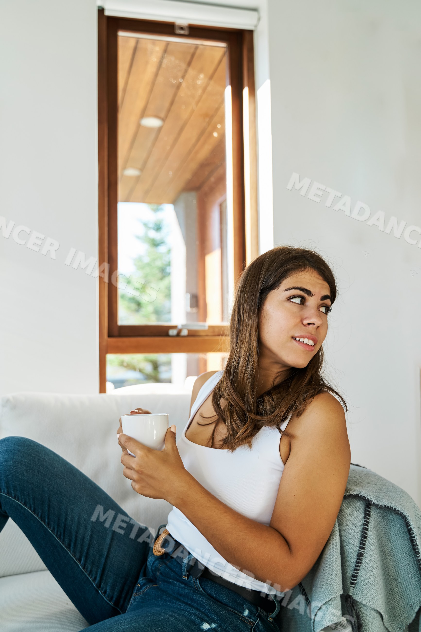 A young white woman relaxes on a couch holding a mug by a window