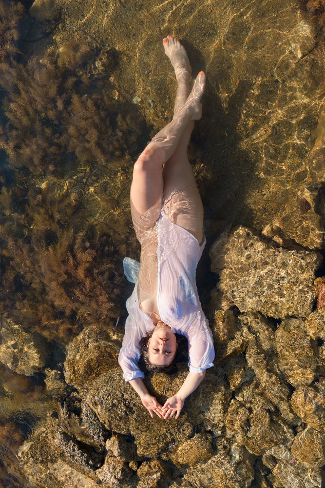 White woman relaxing in shallow water on rocks surrounded by seaweed