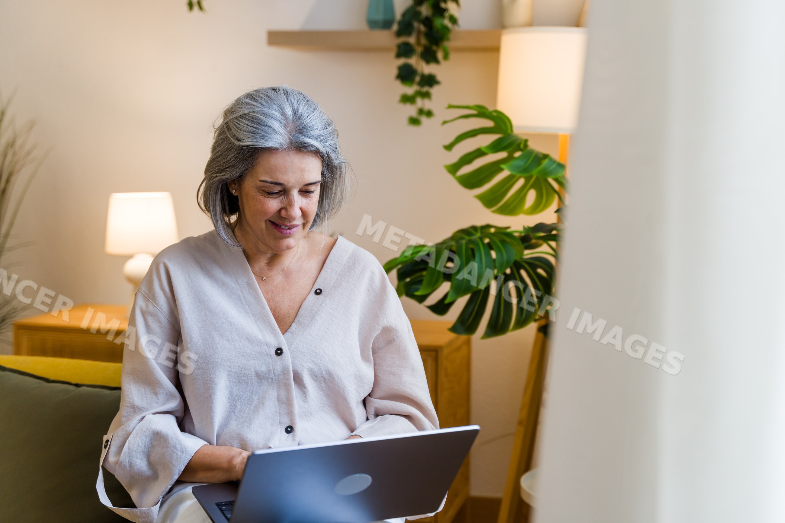White woman smiling while using laptop indoors near plants and lamps