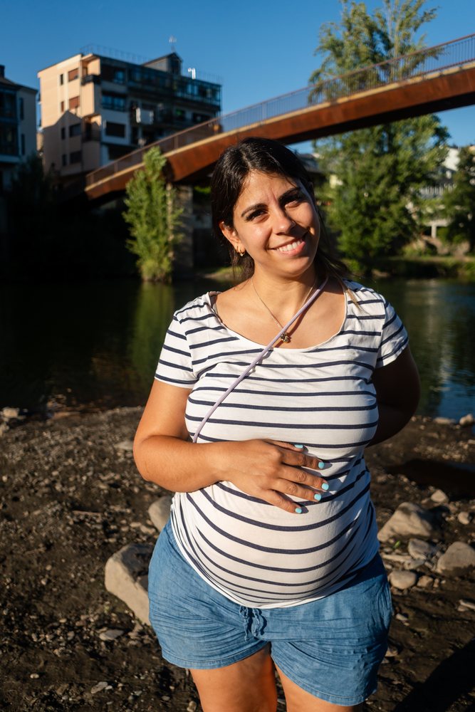 Pregnant woman smiling outdoors by a riverbank with a bridge