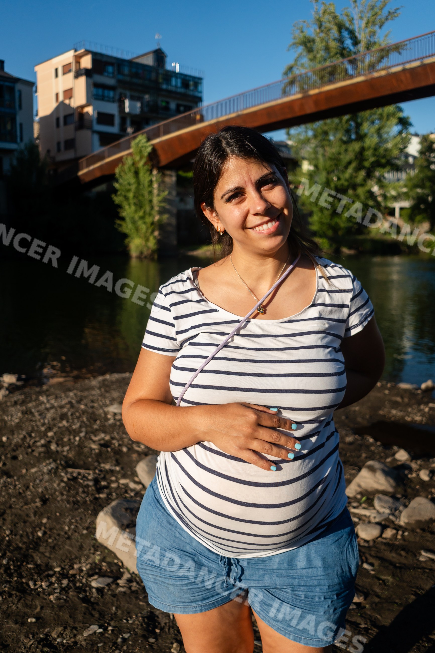 Pregnant woman smiling outdoors by a riverbank with a bridge