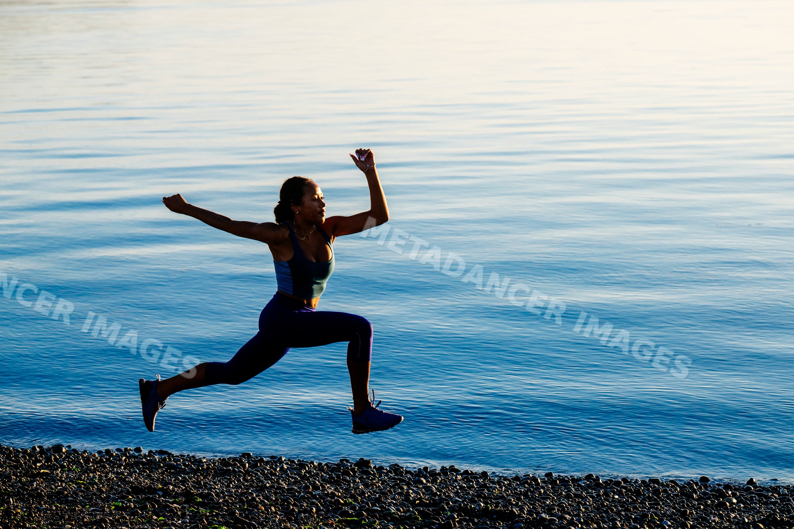 Hispanic woman jumping in athletic pose on pebble beach by lake