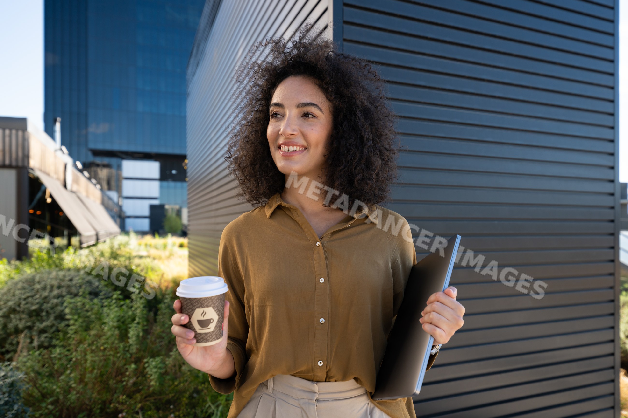 Woman holding coffee cup and laptop outdoors by modern building