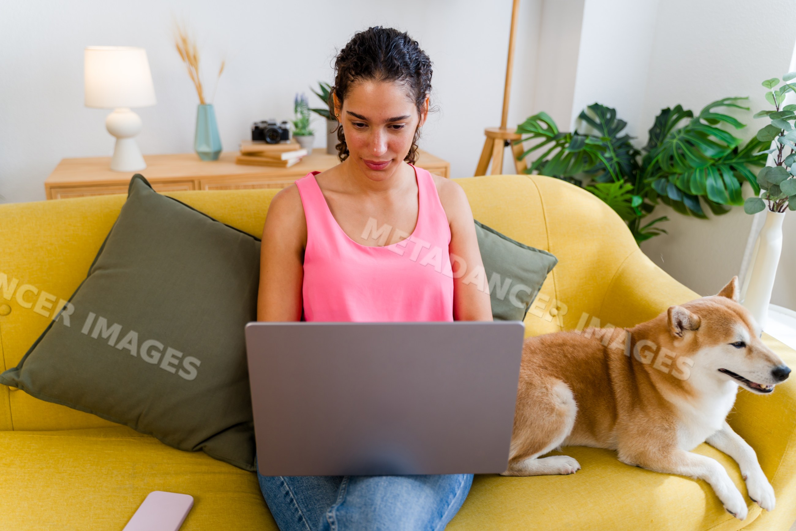 A Hispanic woman works on a laptop sitting on a yellow sofa with her
