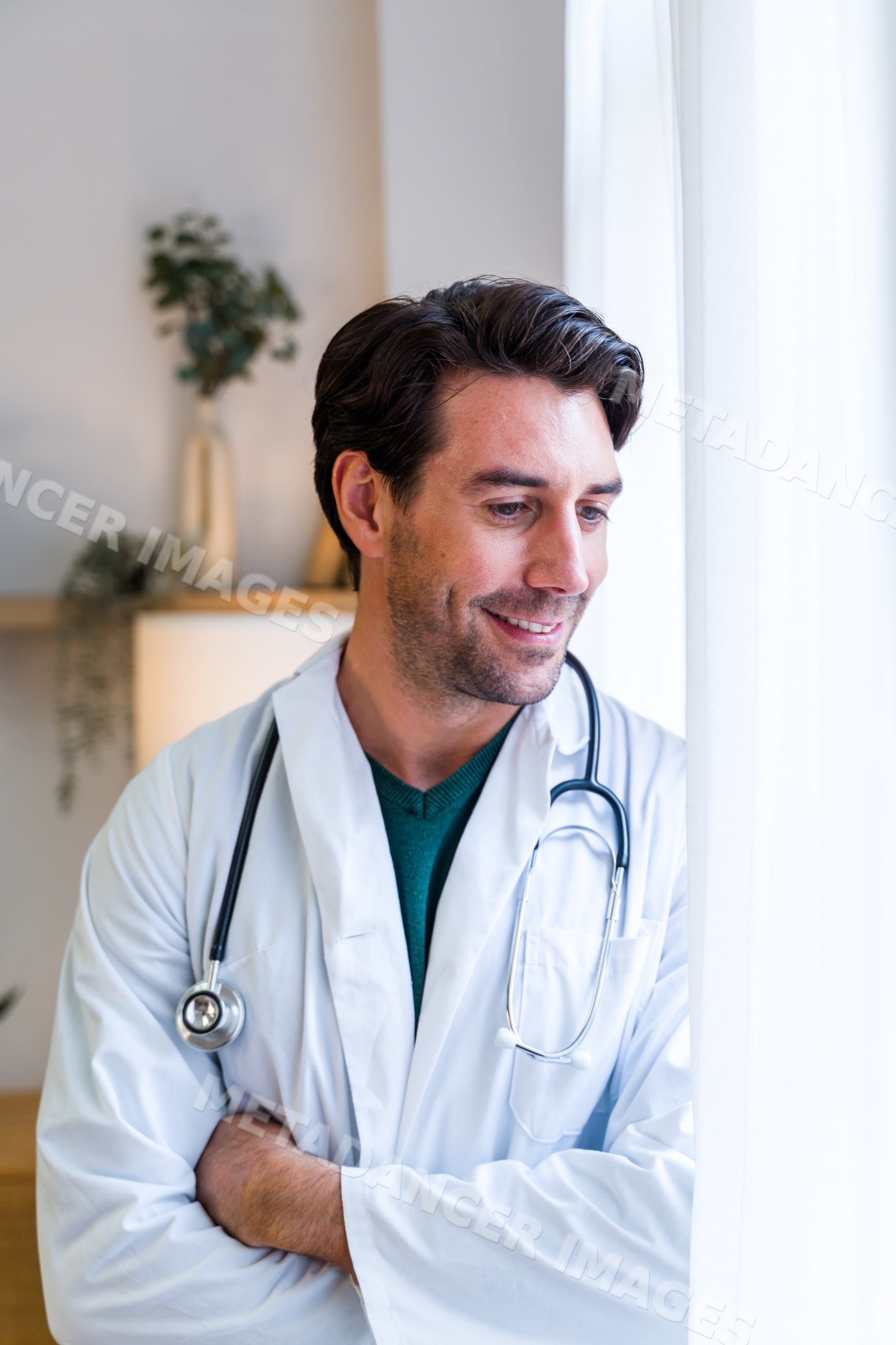 Doctor in white coat with stethoscope looking out window indoors
