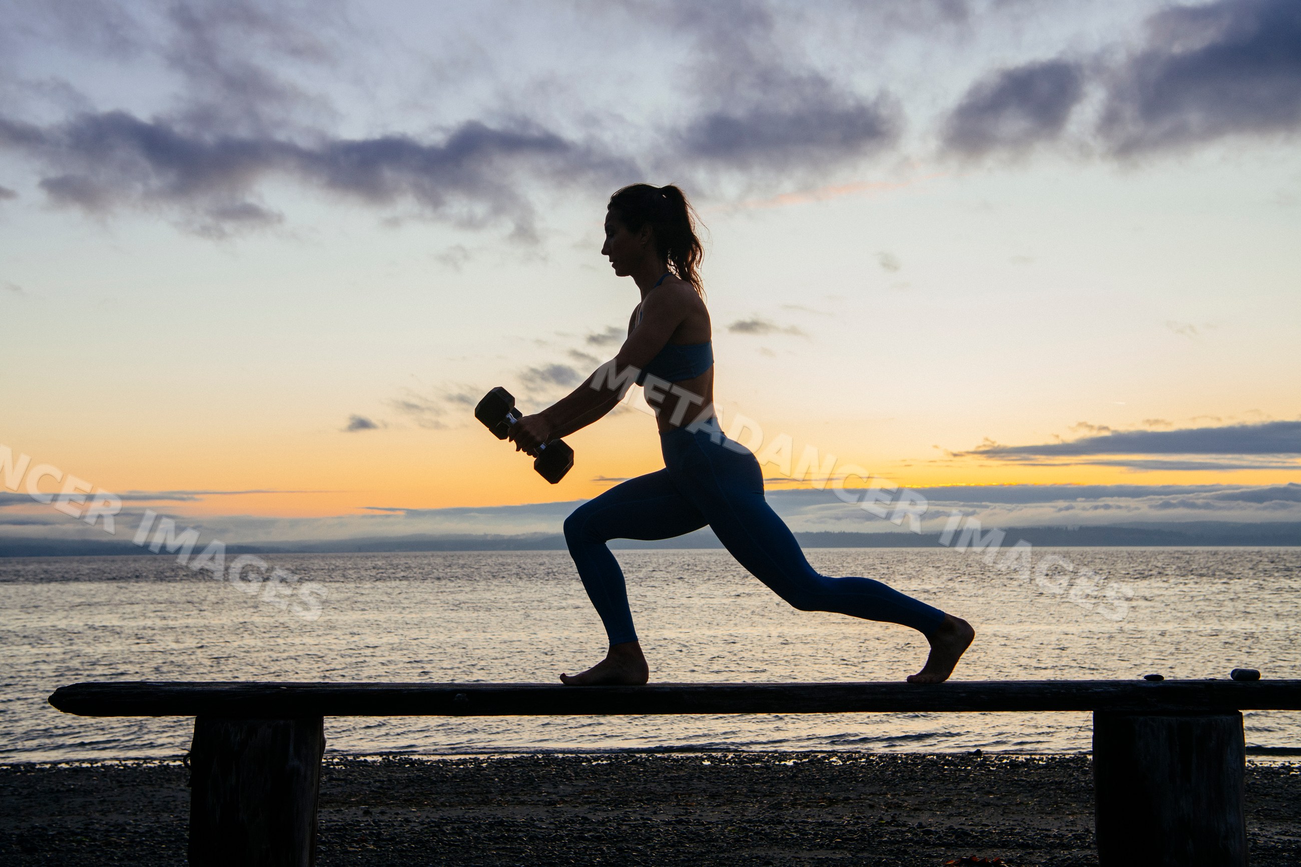 White woman exercising with dumbbells on bench at sunset by water