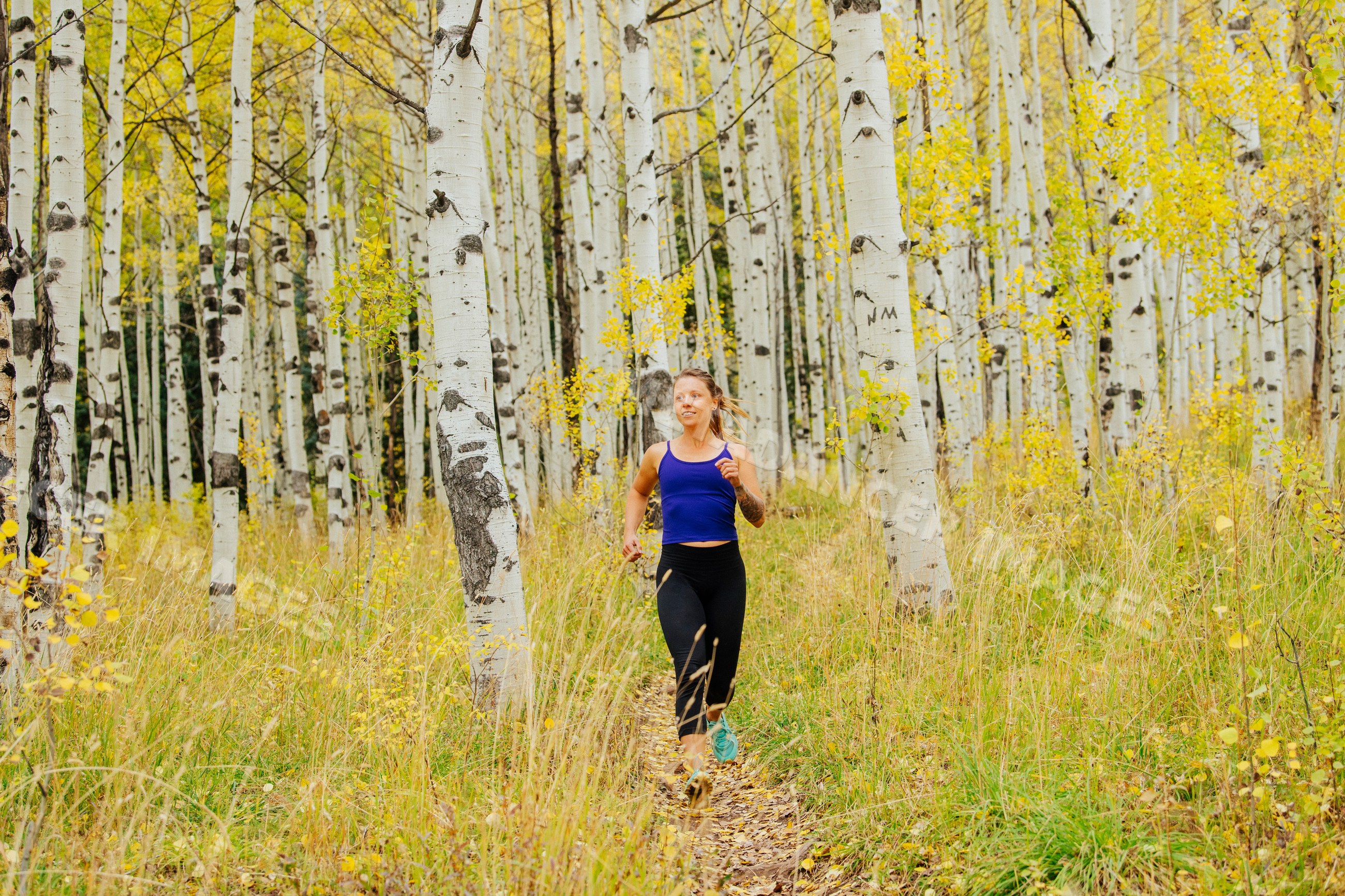 White woman running on trail in birch forest with yellow autumn leaves