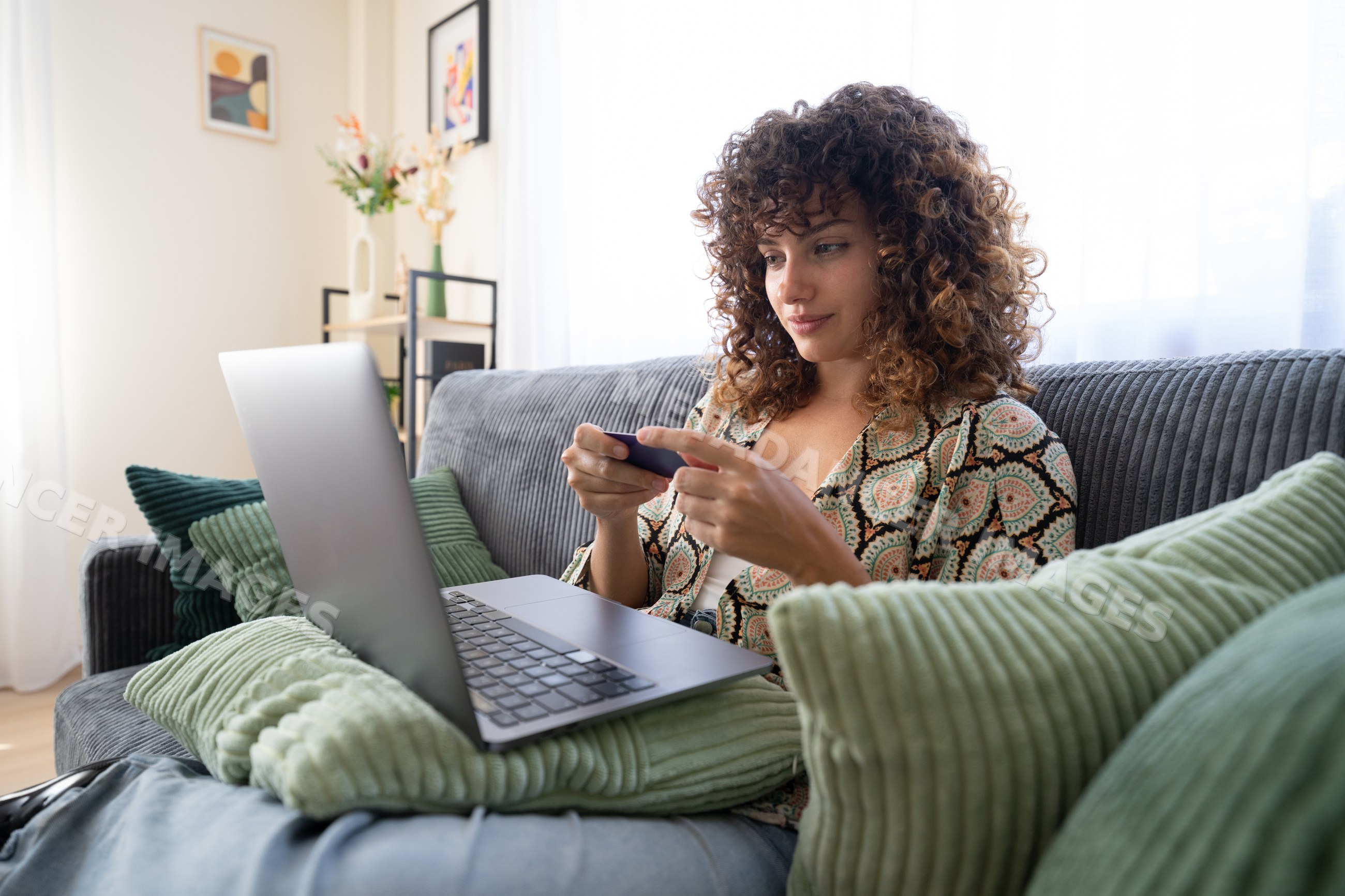 White woman using smartphone on couch with laptop and pillows