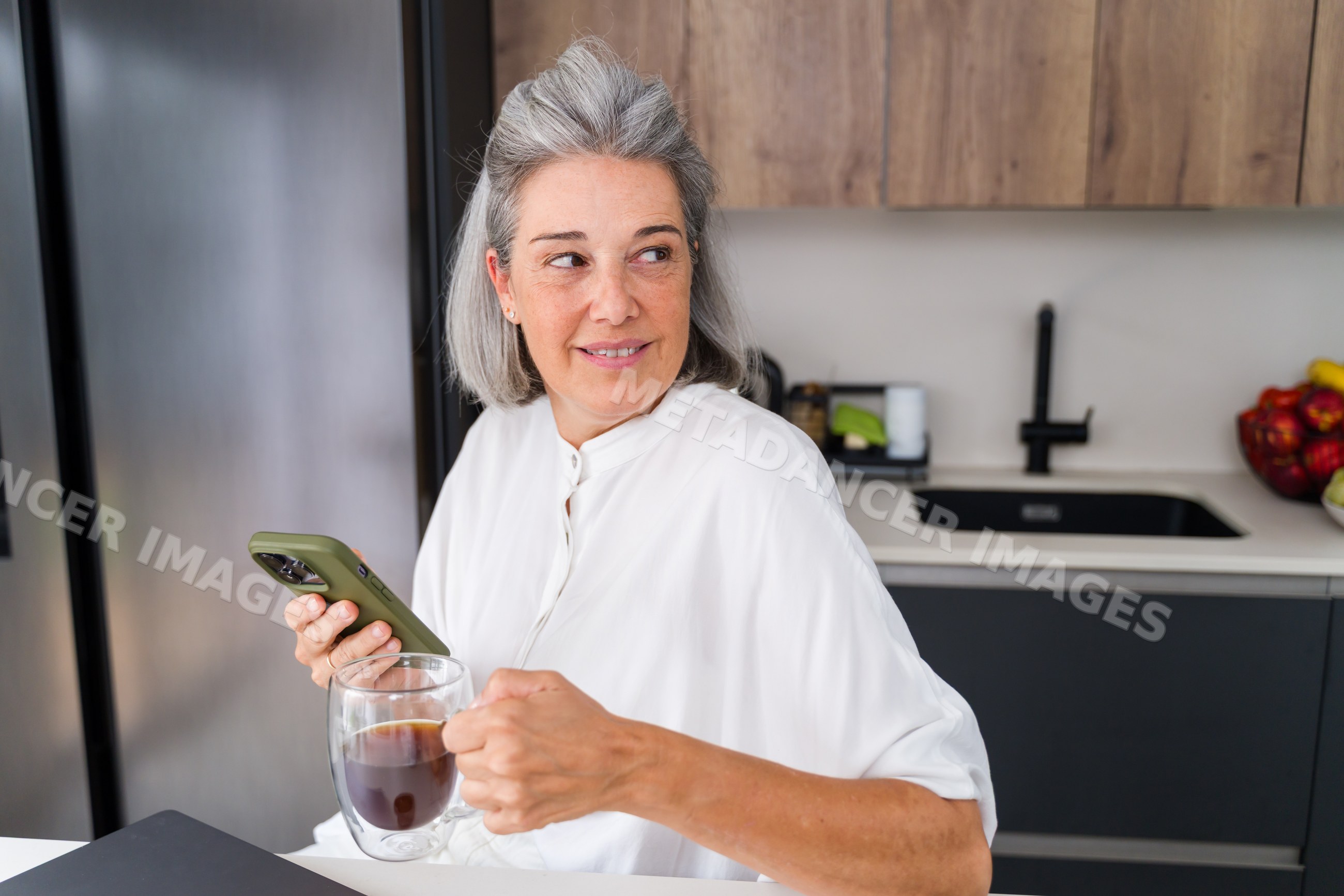 Mature white woman holding smartphone and coffee in modern kitchen