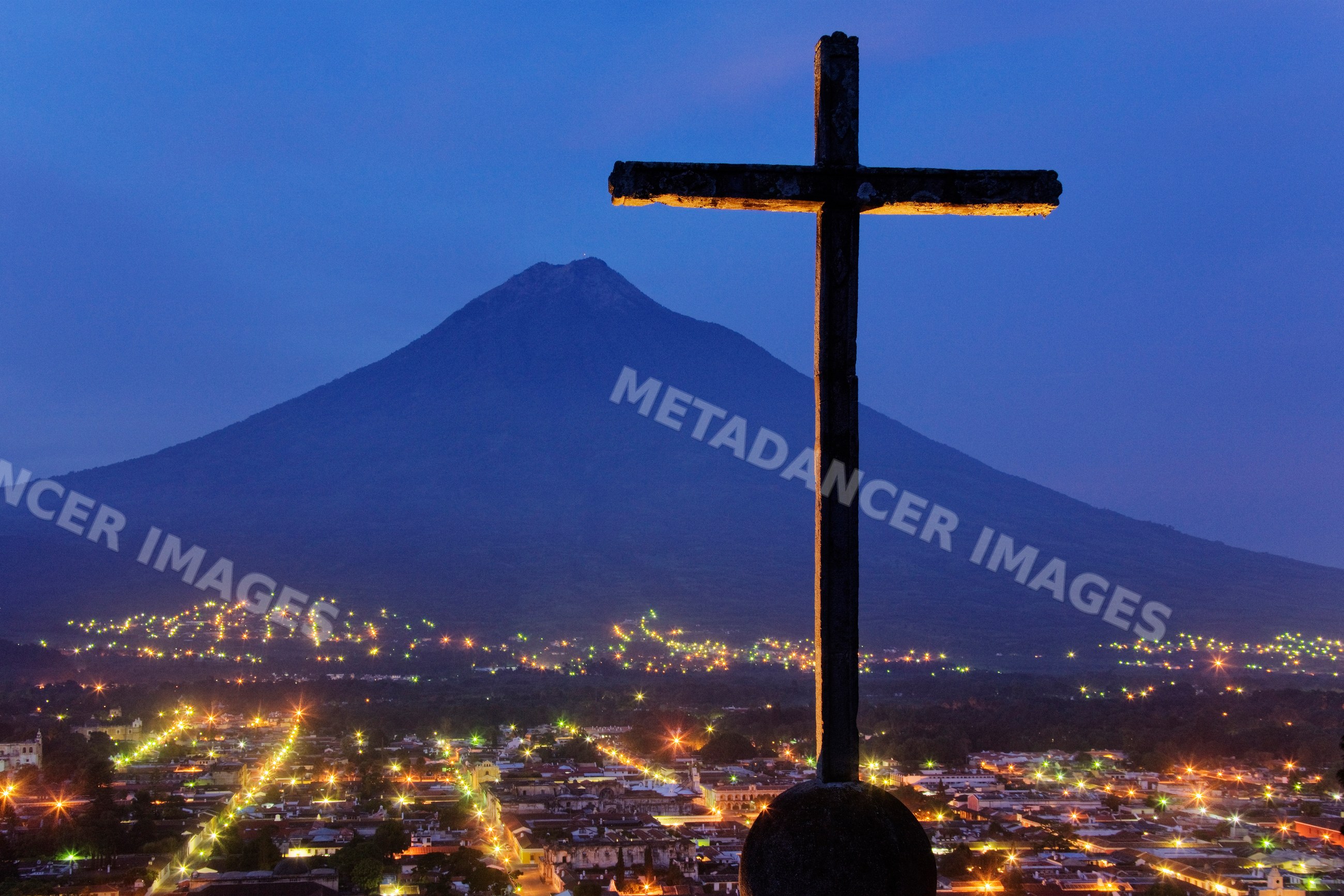 Cross silhouette at volcan agua mountain with city lights at dusk