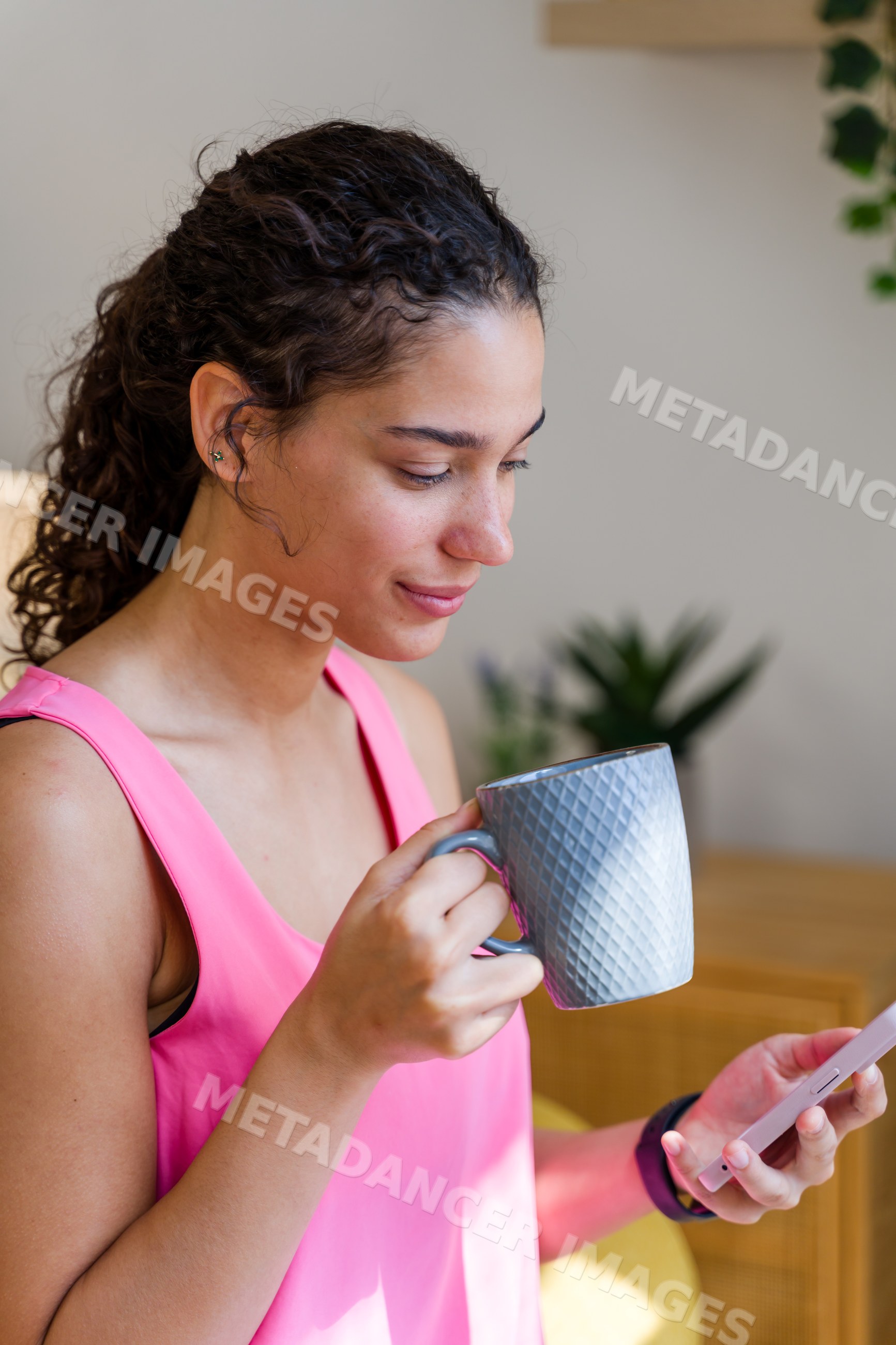 Young Hispanic woman drinking from mug while using smartphone indoors