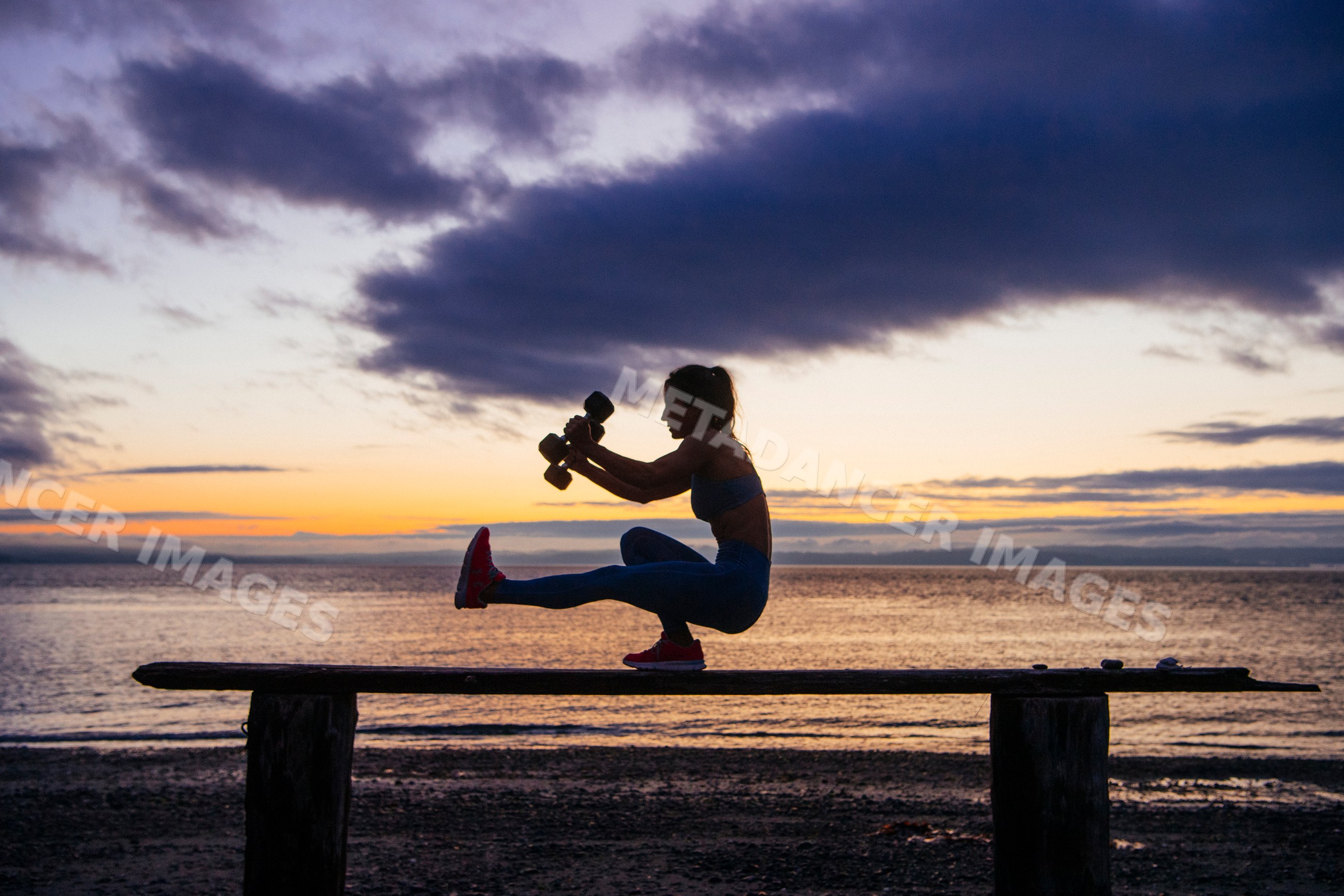 White woman balancing with dumbbells on bench sunset in a beach