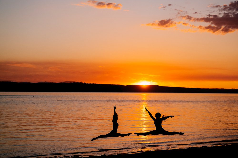 Two white people leap in yoga partner acrobatics by a lake at sunset