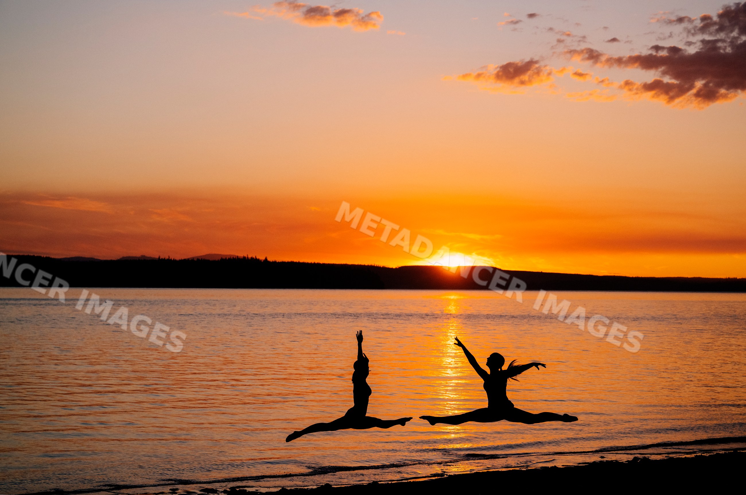Two white people leap in yoga partner acrobatics by a lake at sunset