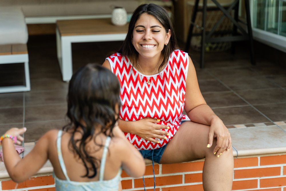 A smiling white woman sits on a brick ledge interacting with a child
