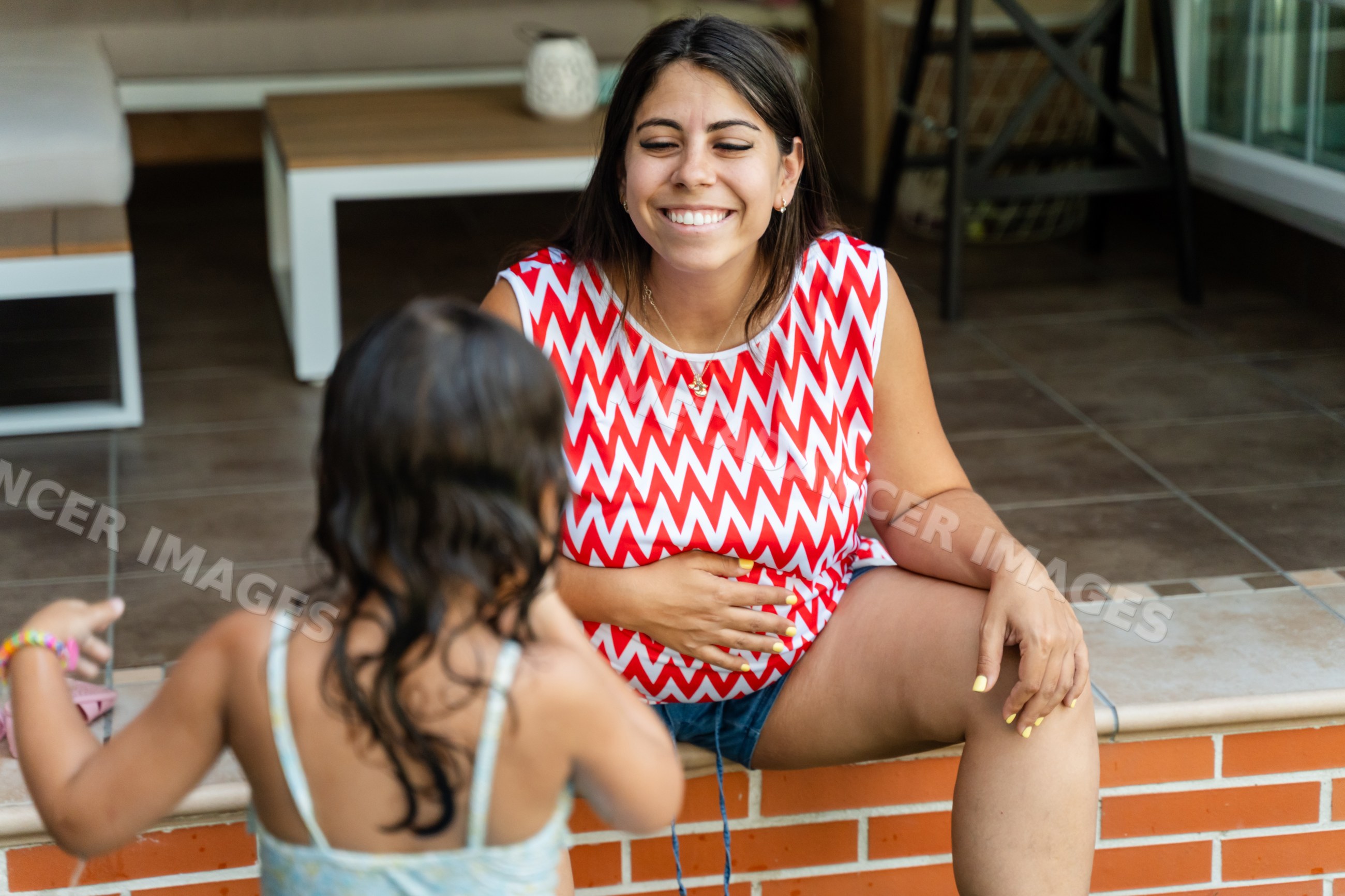 A smiling white woman sits on a brick ledge interacting with a child