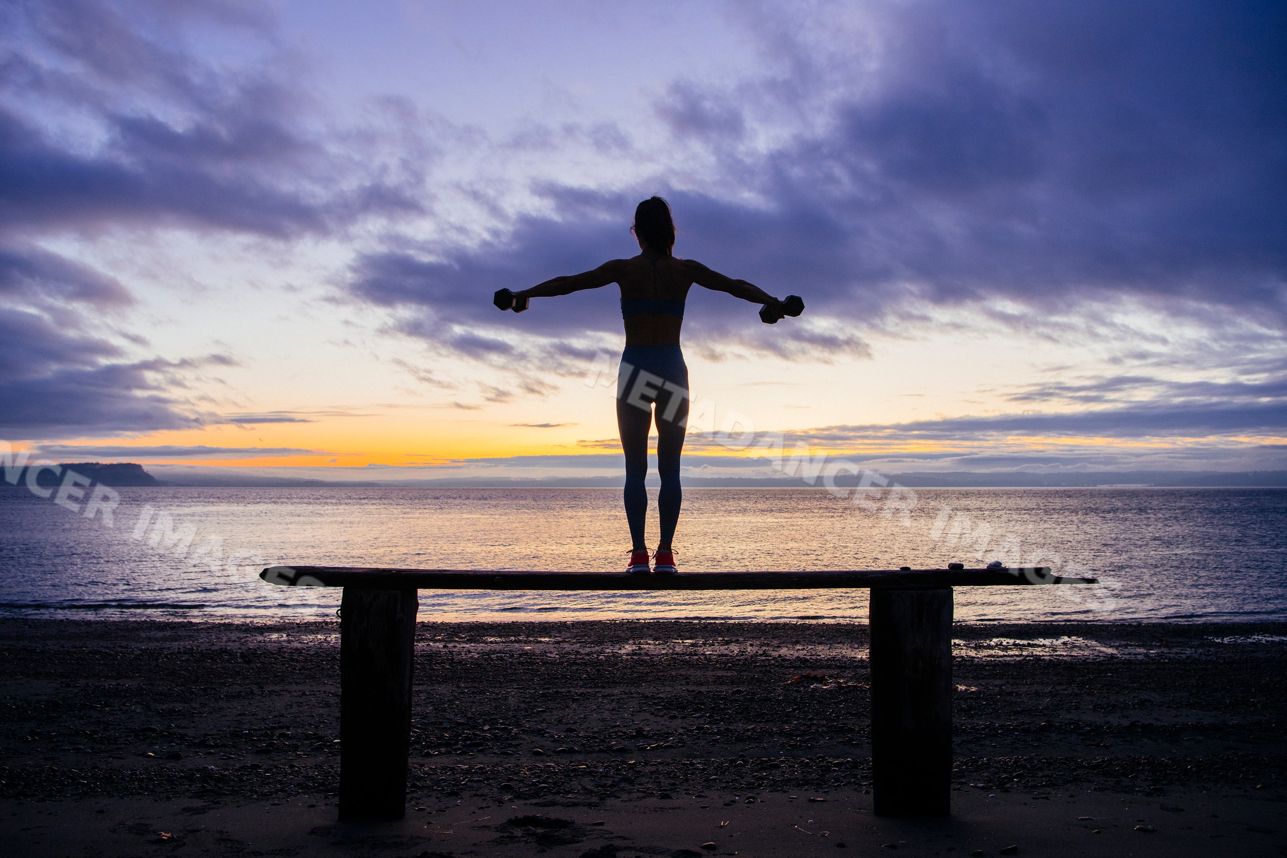 White woman exercising with dumbbells balancing on a beach beam