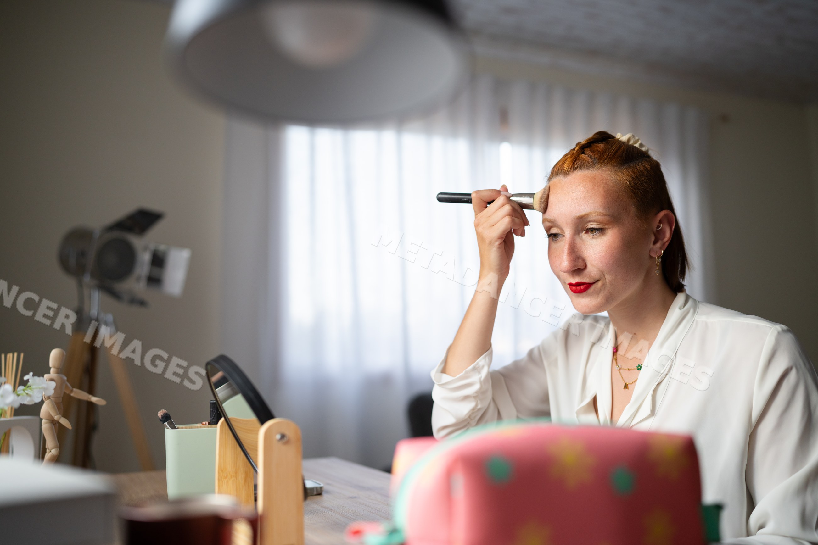 A white woman applies makeup at a vanity in a cozy home setting