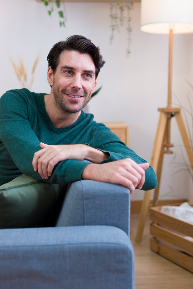White man smiling while leaning on couch in home interior