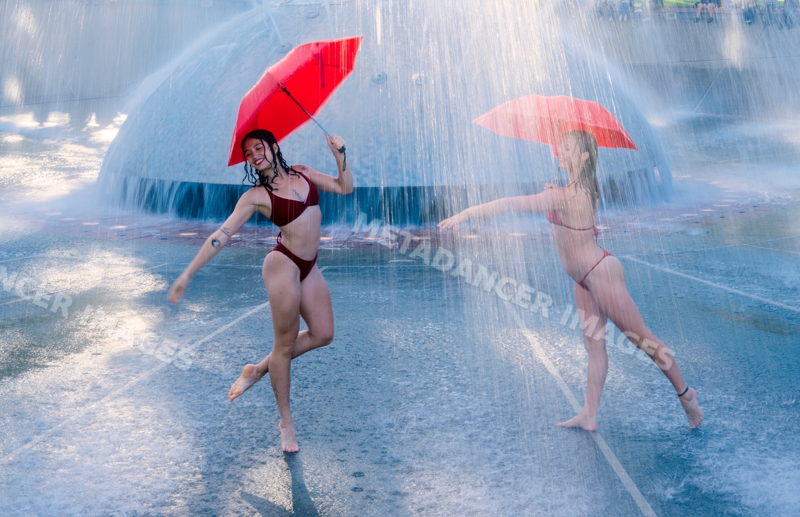 Two white women dancing with red umbrellas under water fountain spray