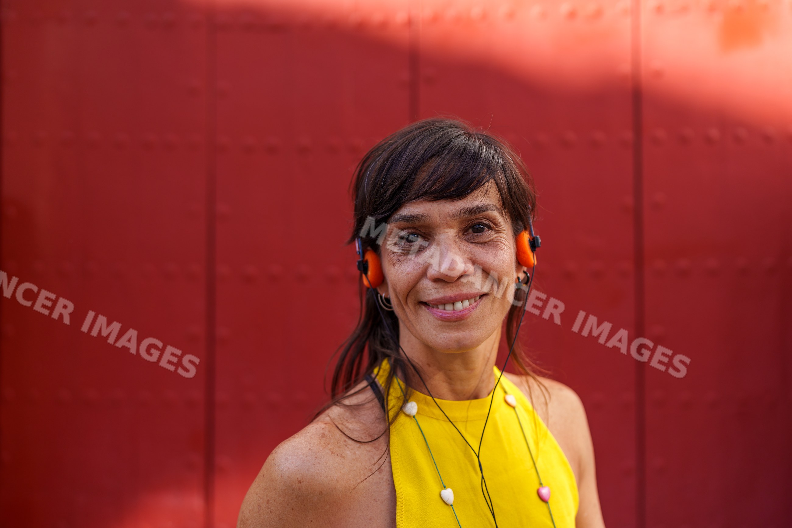 Hispanic woman smiling with orange headphones outside by red wall