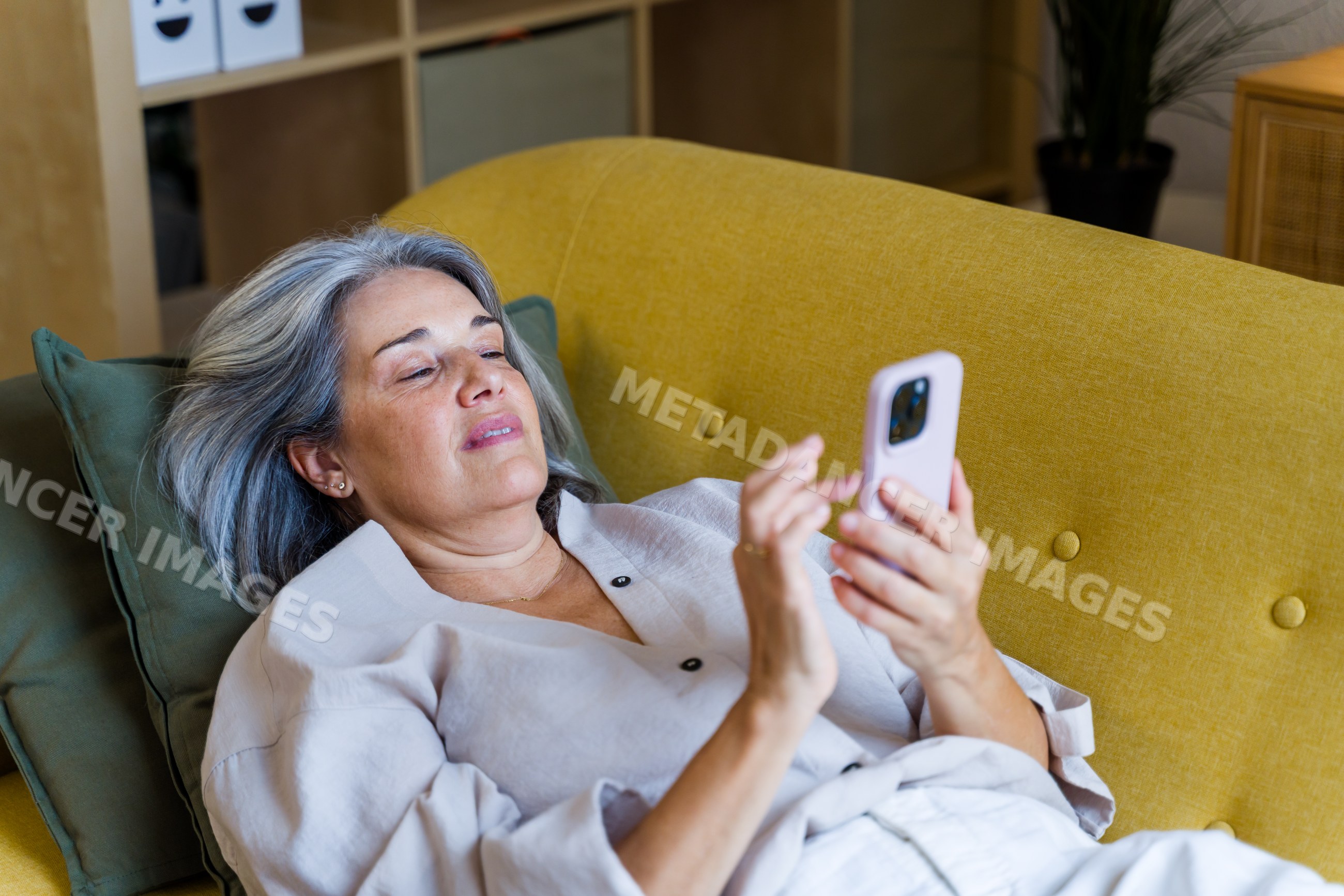 Middle-aged white woman relaxing on yellow couch using smartphone